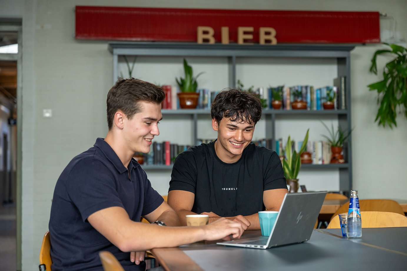 twee mannen werken samen aan een laptop, met koffie en de boekenkast achter zich